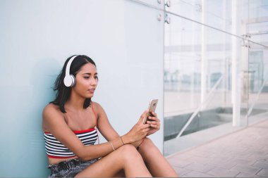 Düşünceli etnik kadın, kaldırımda otururken ve caddede cam duvara yaslanırken cep telefonuyla şarkılar dinliyor.