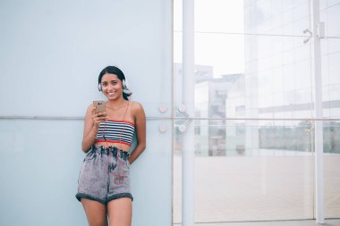 Yazlık giysiler içinde genç etnik bayan hipster sokakta duruyor ve cam duvara yaslanıp kameraya bakarken cep telefonundan müzik listesinden şarkılar dinliyor.