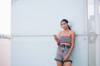 Moda kıyafetli genç bir bayan akıllı telefondan şarkılar dinliyor ve modern binanın cam duvarına yaslanıp sokakta dinleniyor ve kameraya bakıyor.