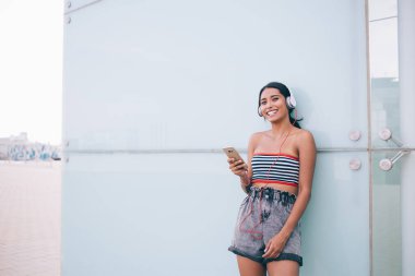 İyi ki yaz kıyafetli bir bayan hippi akıllı telefonuyla caddede duruyor ve cam duvara yaslanıp kameraya bakarken kulaklıkla şarkılar dinliyor.