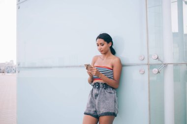 Genç, sıska esmer, yazın üstte ve kısa taramalı akıllı telefonlu şehir caddesindeki modern binanın duvarında dikilirken