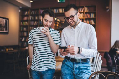 Akıllı telefonları karıştıran ve sıcak içecekler içen neşeli gençler kütüphanedeki serbest meslek sırasında masaya yaslanıp dinleniyorlar.