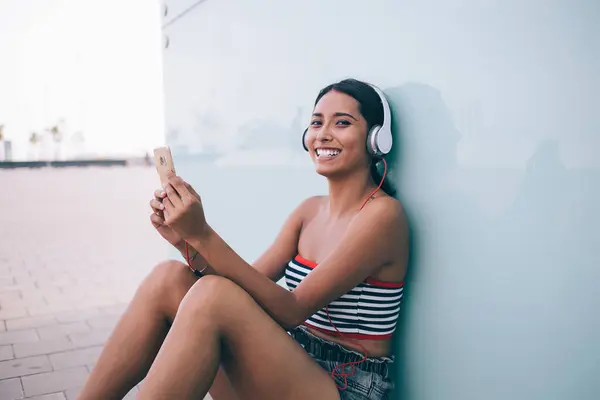 Cheerful female hipster in summer outfit sitting on sidewalk and listening to songs in headphones via mobile phone while leaning on glass wall and looking at camera