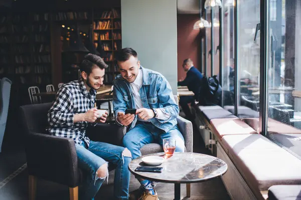 Joyful young male friends in stylish casual clothing watching media content on mobile phone while sitting in modern cafe with tea and coffee