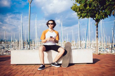 Genç, kıvırcık saçlı, güneş gözlüklü ve sıradan kıyafetli bir adam. Barcelona 'daki güzel rıhtımda kayalık çitlerde otururken cep telefonuyla sohbet ediyor. 