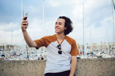 Neşeli kıvırcık saçlı erkek gezgin gülümseyerek gülümserken cep telefonuyla barselona arka planında renkli iskele olan çitlere yaslanarak selfie çekiyor. 