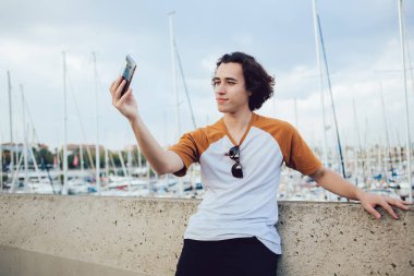 Genç erkek blogger 20 'li yaşlarda medya paylaşımı için medya içeriği yaratmak için ön kameraya bakarken, genç hippi arka planda kendini çekerken selfie görüntülerine tıklıyor.