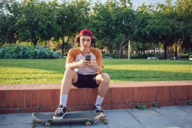 Havalı giyinen siyah saçlı genç adam spor ayakkabı giyer ve kırmızı şapkalı kaykaylı bacaklarıyla çitlerde oturur ve Barselona 'da cep telefonu kullanır. 