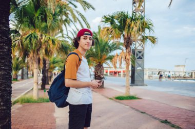 Sıradan giyinen, sırt çantalı, şapkalı, gülümseyen ve elinde cep telefonuyla Barcelona 'da egzotik avuçlarıyla kameraya bakan içerikli erkek hipsterin yan görüntüsü. 