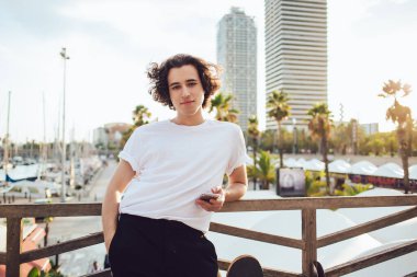 Barcelona 'da cep telefonuyla ahşap çitlere yaslanırken kameraya bakan, modaya uygun giyinen iri yapılı genç adam.