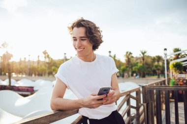 Beyaz gömlekli, siyah pantolonlu, çitlere yaslanmış gülümseyen genç bir adam. Güneşli Barcelona 'da telefonu elinde tutan dişlek bir gülümsemeyle başka tarafa bakıyor.