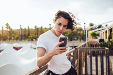 Yakışıklı, kıvırcık saçlı, gündelik giyinmiş, Barcelona 'da ahşap çitlere yaslanırken cep telefonu izleyen bir adam.