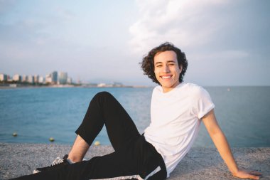 Beyaz tişörtlü genç adam taştan çitlerin üzerinde oturuyor şehir ve deniz manzarasına karşı kameraya gülümserken güneşleniyor.