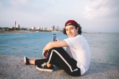 Beyaz tişörtlü hippi kıyı şeridinin çitlerinde cep telefonu kullanarak kameraya gülümseyen ve fotoğraf çeken adamın yan görüntüsü.