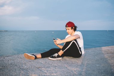 Kırmızı şapkalı düşünceli genç adam gün batımında temiz suya ve bulutlu gökyüzüne karşı taş çitlerde serinlerken akıllı telefonuyla mesaj atıyor.