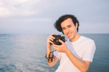 Deniz ve bulutlu gökyüzünün arka planında duran mutlu genç adam gün batımının fotoğrafını çekiyor ve yaz tatilinin tadını çıkarırken gülümsüyor.