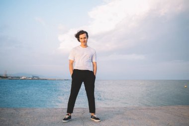 Neşeli hippi adam, gündelik giysiler içinde taş çitin üzerinde el ele tutuşmuş ve denizde vakit geçirirken kameraya bakıyor.