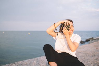 Deniz kenarındaki arka planda oturan yeniyetme erkek, taş çitlerde oturuyor ve günbatımının fotoğraflarını çekiyor.