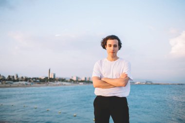 Beyaz tişörtlü düşünceli bir adam taş çitin üzerinde el ele tutuşup kameraya bakarken hayal kuruyor ve gezinin tadını çıkarıyor.