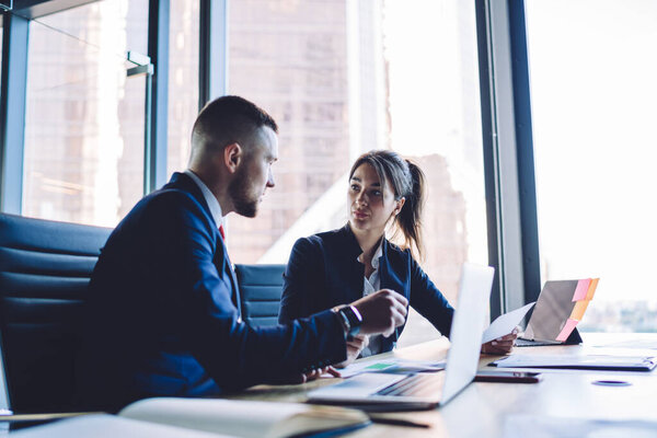 Caucasian persons discussing revenue capital during brainstorming on business startup project, formally dressed male and female colleagues cooperating at table desktop with laptop and documents