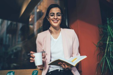 Eğitim kitabı ve kahvesi olan zeki bir bayan öğrenci kameraya gülümseyecek. Optik gözlüklü neşeli İspanyol kadının yarım boy portresi kaldırımda bilgi öğreniyor.