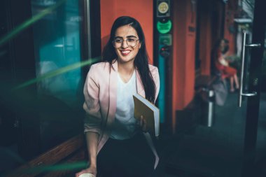 Görme koruması için gözlüklü mutlu İspanyol bayan öğrenci edebiyat hobisi için zaman harcıyor, şehir ortamında çok satan kitabı olan neşeli Gürcü kadın gülümsüyor.