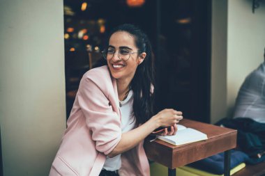 Şık klasik gözlüklü mutlu bayan öğrenci sokak kafeteryasında eğitim kitaplarıyla vakit geçiriyor, gözlüklü neşeli Gürcü kadın kafe terasında gülümsüyor.