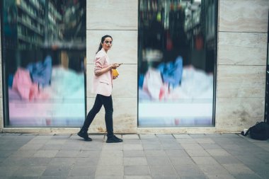 Eğitimli not defteri ve akıllı telefon aleti olan modaya uygun giyinmiş bir kız öğrencinin şehir merkezinde yürüyen ve kameraya bakan cep telefonlu İspanyol bir kadının tam boy portresi.