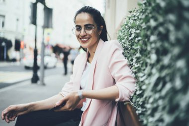 Şehir bankında dinlenirken kameraya gülümseyen moda gözlüklü komik kadın blogcu portresi, akıllı telefon teknolojisiyle şehirde keyif çatan mutlu İspanyol kadın.