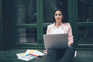 Laptop bilgisayarlı düşünceli bayan öğrenci araştırma sırasında grafik grafikleri hakkında düşünüyor ve şehir ortamında öğreniyor, gözlüklü İspanyol kadın bilgi grafikleri üzerinde uzaktan çalışıyor.