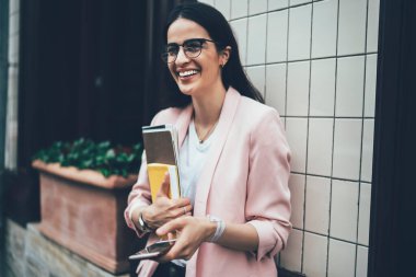 Moda gözlüklü neşeli hippi öğrenci elinde ders kitabı ve cep telefonu zımbırtısı, mutlu kadın blogcu şehir ortamında duruyor ve akıllı telefon cihazıyla sosyal ağ kurarken gülüyor.