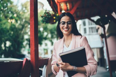 Şehir merkezinde poz veren neşeli kadın gazetecinin yarım boy portresi, klasik gözlüklü başarılı Gürcü kadın şehirde boş zamanlarında kameraya gülümsüyor.