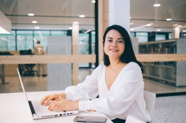 Masada dizüstü bilgisayarla çalışan, klavyede daktilo kullanan ve ofisteki cam duvara gülümseyen kendine güvenen modacı bir bayan.
