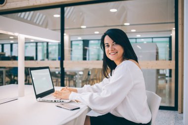 Modern ofiste sandalyede oturan ve kameraya gülümserken boş ekranla dizüstü bilgisayarda çalışan beyaz bluzlu mutlu genç bayan. 