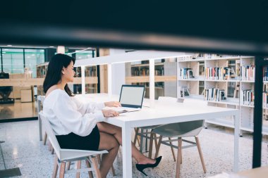 Masada oturmuş, sınava hazırlanırken dizüstü bilgisayar kullanan siyah saçlı, şık bayan öğrencinin yan görüntüsü. 