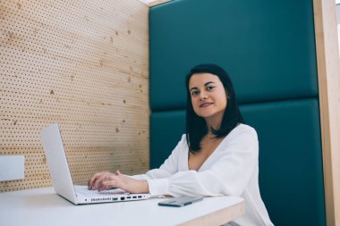 Barcelona 'da modern bir kafede dizüstü bilgisayarda gezinirken gülümseyen ve kameraya bakan beyaz bluzlu muhteşem siyah saçlı serbest kadın.