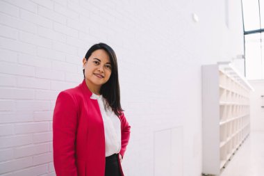 Resmi giyinmiş, renkli pembe ceketli, zarif siyah saçlı kadın Barcelona 'daki beyaz duvarın önünde gülümseyip kameraya bakıyor.