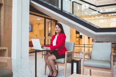 Modern ofiste çalışırken ve kameraya bakarken resmi giyinmiş siyah saçlı, etnik bir kadın masada oturuyor ve netbook 'ta sörf yapıyor.