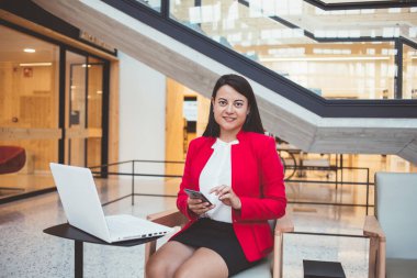 Genç Latin kökenli, siyah saçlı, cep telefonuyla yazı yazan, modern ofiste çalışırken masasında beyaz dizüstü bilgisayarıyla oturan bir kadın.