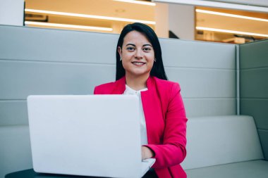 Resmi giysiler içinde genç İspanyol bayan kanepeye oturmuş, bilgisayar başında, modern ofiste proje üzerinde çalışıyor ve kameraya bakıyor.