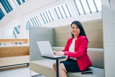 Beyaz bluzlu ve siyah etekli etnik kadın koltukta oturuyor cep telefonu rehberi ile ve parlak ofiste çalışmanın tadını çıkarıyor.