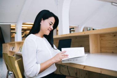 Modern kütüphanede güneşli bir günde masa başında otururken keyifli bir şekilde kitap okuyan neşeli genç bayanın yan görüntüsü.