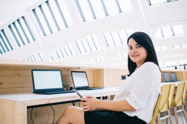 Resmi giysiler içinde gülümseyen bir kadının yan görüntüsü. Akıllı telefonuyla mesajlaşıyor ve kameraya bakarken sandalyede oturuyor.