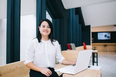 Modern açık uzay kütüphanesinde beyaz bilgisayar projesinde çalışan esmer kadın gülümseyip kameraya bakıyor.