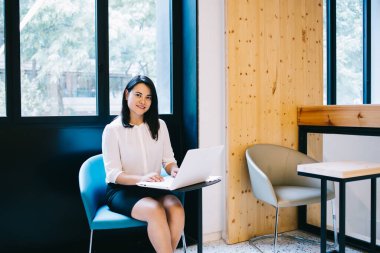 Netbook 'ta yazan ve başlangıç stratejisi üzerinde çalışan kadın bir uzman rahat koltukta oturmuş kameraya bakıyor.