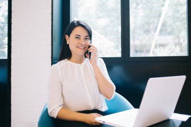 Zarif kıyafetler giymiş, masada dizüstü bilgisayarla oturup kameraya bakarken telefonla iletişim kuran genç gülümseyen yönetici asistanı.