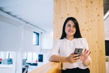 İyi giyimli, gülümseyen iş kadını akıllı telefonu kaydırıyor ve geniş ofis işyerinde kameraya bakarken korkuluklara yaslanıyor.