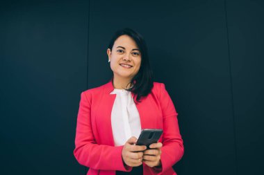 Zarif bir ceket giyen, akıllı telefondan gülümseyen ve internette sörf yapan bayan patron ofis mavi duvarının arkasında kameraya bakıyor.