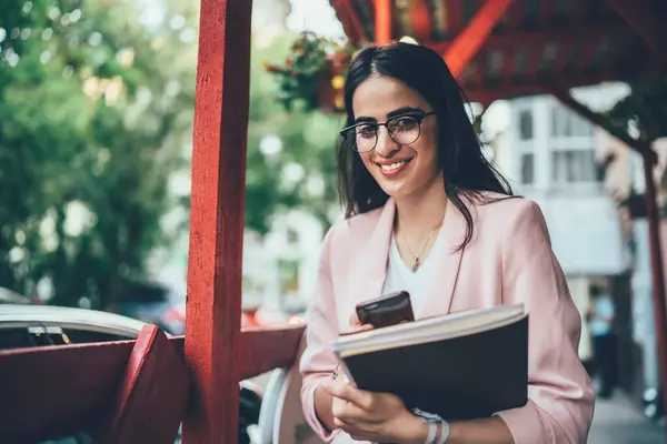 Half length portrait of cheerful influence blogger with modern cellphone device and textbook spending leisure for creating content publication, sincerely woman in trendy glasses holding equipment