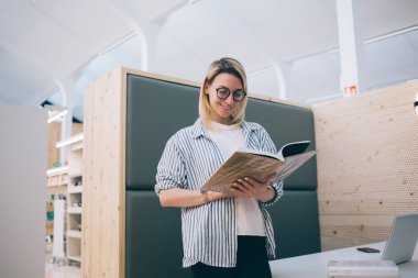 Barselona 'daki çağdaş üniversite kütüphanesinde dizüstü bilgisayarla oturmuş gülümseyen ve kitap okuyan gözlüklü mutlu genç öğrenci.